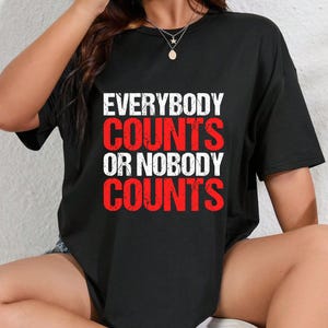 Könnte beinhalten: Ein schwarzes kurzärmeliges T-Shirt mit dem Text "EVERYBODY COUNTS OR NOBODY COUNTS" in einem Used-Look-Druck. Die Wörter "EVERYBODY" und "OR NOBODY" sind weiß, während "COUNTS" zweimal in einer fetten, leuchtend roten Schrift erscheint.
