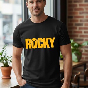 May include: A black short-sleeved t-shirt featuring the word ROCKY in bold yellow capital letters across the chest, with a small silhouette of a person with arms raised positioned inside the letter O of the word.