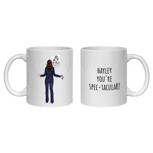 Könnte beinhalten: Ein weißer Keramikbecher mit einer Grafik einer Person im dunklen Anzug vor einem Sehtest-Diagramm und dem Text "HAYLEY YOU'RE SPEC-TACULAR!" in Schwarz auf der gegenüberliegenden Seite, mit einem Henkel und einem glatten zylindrischen Körper.