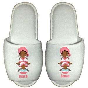 May include: White spa slippers with a cartoon illustration of a woman with brown skin, wearing a pink robe and a pink towel on her head, soaking her feet in a blue bowl. The slippers have the name "Grace" printed on the bottom.