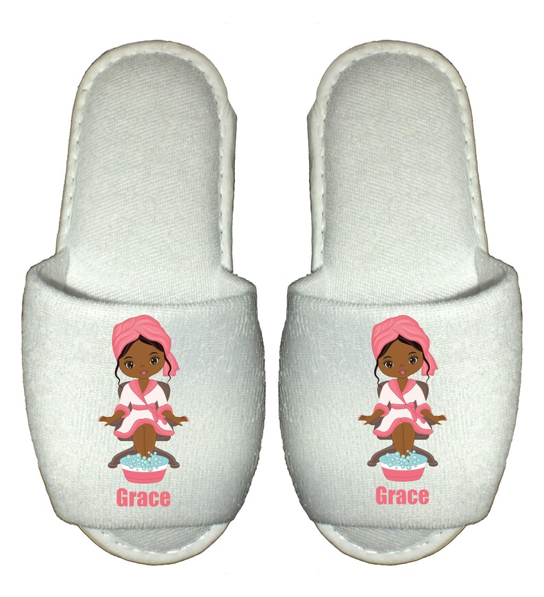 May include: White spa slippers with a cartoon illustration of a woman with brown skin, wearing a pink robe and a pink towel on her head, soaking her feet in a blue bowl. The slippers have the name "Grace" printed on the bottom.