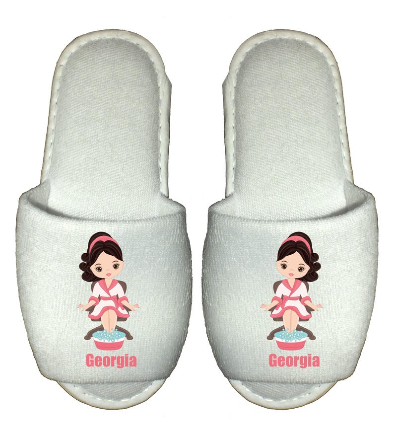 May include: White spa slippers with a cartoon illustration of a woman getting a pedicure. The slippers have the name "Georgia" printed on them.
