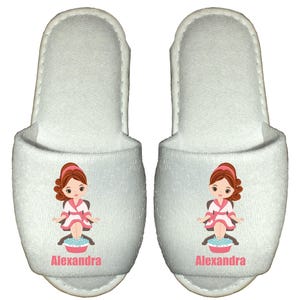 May include: White spa slippers with a cartoon illustration of a woman in a robe with her feet in a bowl of water. The slippers are personalized with the name "Alexandra".