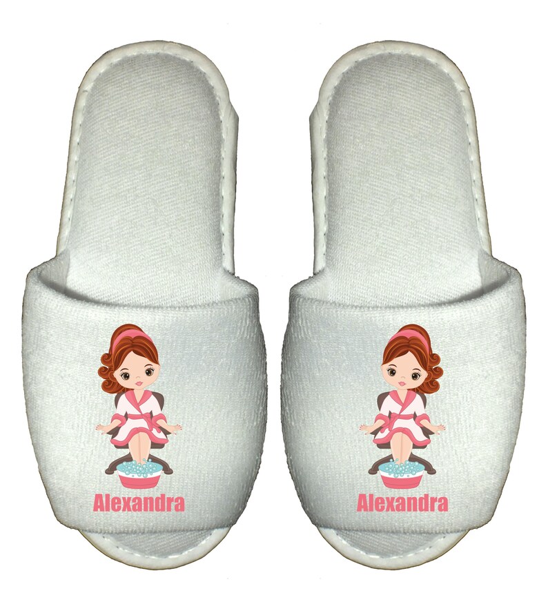 May include: White spa slippers with a cartoon illustration of a woman in a robe with her feet in a bowl of water. The slippers are personalized with the name "Alexandra".