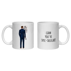 May include: Two white ceramic mugs. One mug features an illustration of a person in a dark blue suit standing before an eye chart, holding glasses. The other mug displays the black text: LIAM YOU'RE SPEC-TACULAR! The mugs have curved handles.
