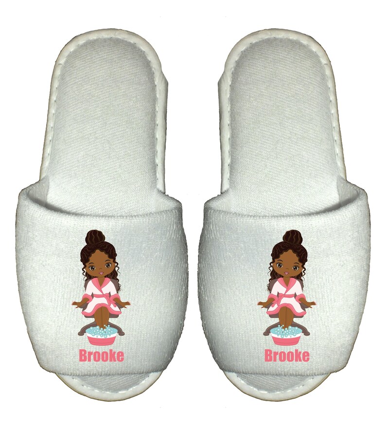 May include: White spa slippers with a cartoon illustration of a girl with brown skin and hair, wearing a pink robe and sitting in a foot bath. The slippers have the name "Brooke" printed on them.