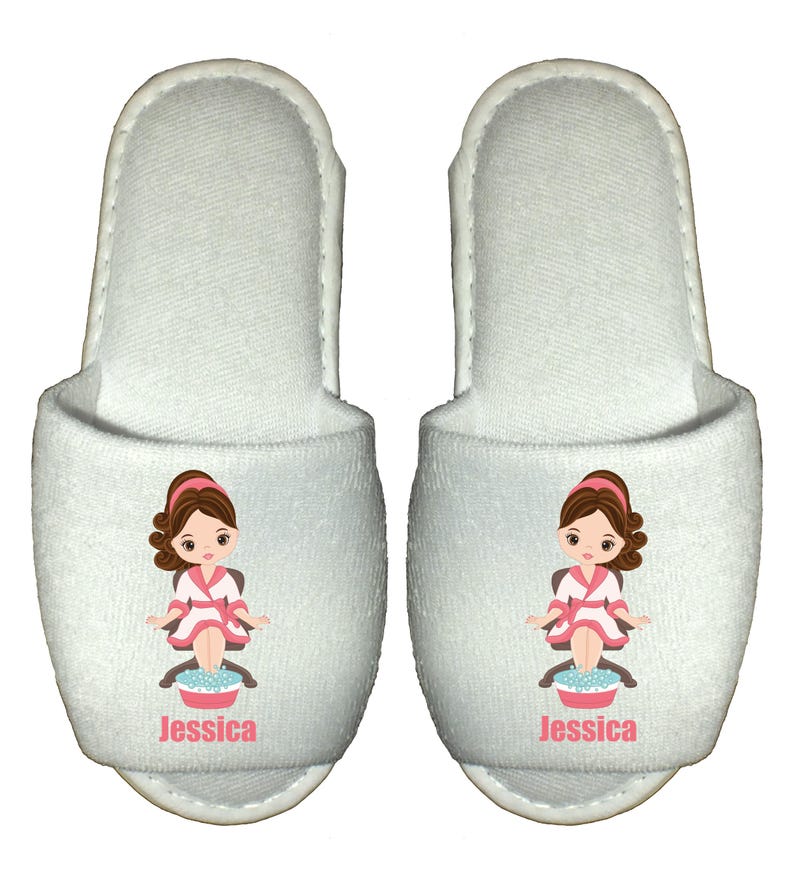 May include: Pair of white spa slippers with a cartoon illustration of a woman getting a foot massage. The slippers have the name "Jessica" printed on them.