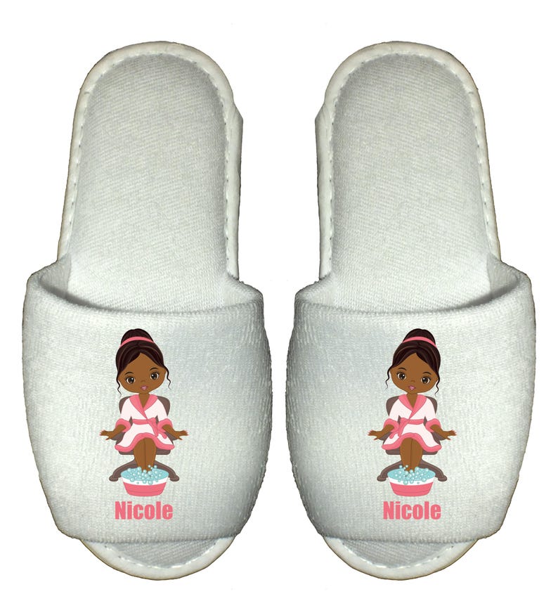 May include: White spa slippers with a cartoon illustration of a woman in a pink robe getting a pedicure. The slippers are personalized with the name "Nicole".