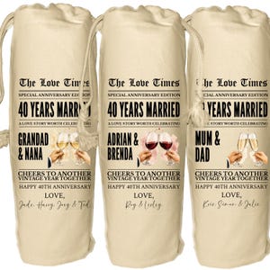 May include: Three beige fabric wine bottle bags with drawstring closures, each printed with 'The Love Times' newspaper-style text, '40 Years Married', and personalized names. The design includes illustrations of hands clinking wine glasses.