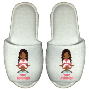 May include: Pair of white spa slippers with a cartoon illustration of a girl in a pink robe with brown hair and skin, soaking her feet in a basin of water. The slippers have the name "Ashleigh" printed on the front.