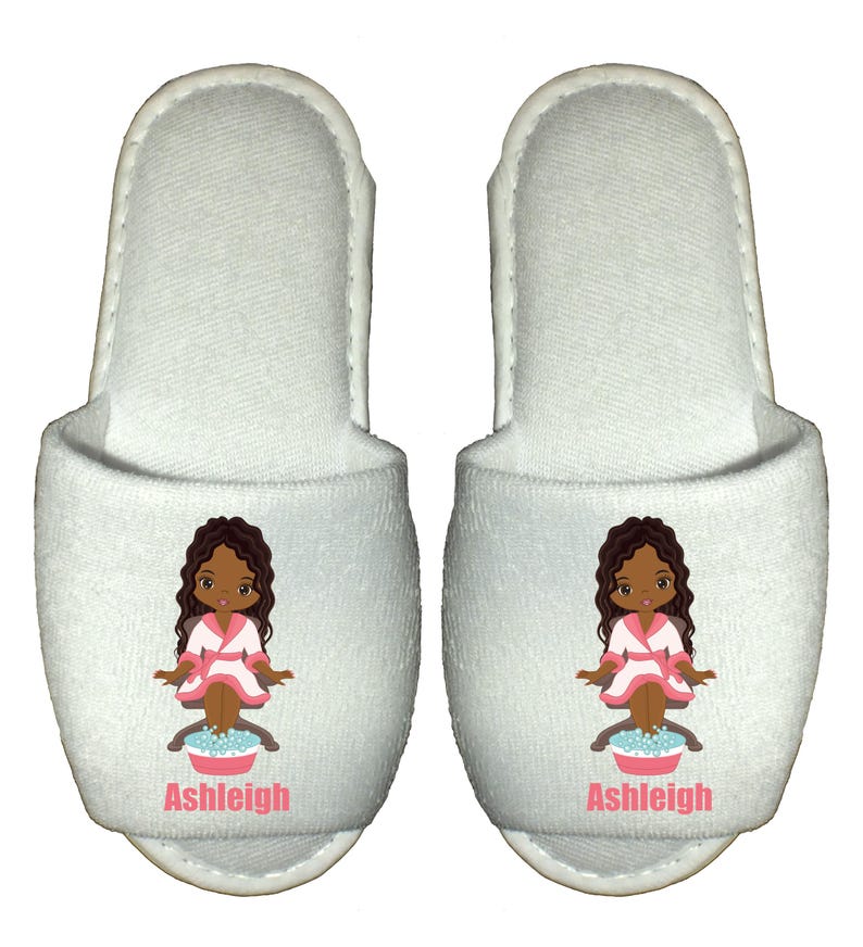May include: Pair of white spa slippers with a cartoon illustration of a girl in a pink robe with brown hair and skin, soaking her feet in a basin of water. The slippers have the name "Ashleigh" printed on the front.