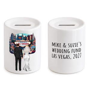 Könnte beinhalten: Zwei weiße Keramik-Spardosen mit einem Schlitz auf der Oberseite. Eine Dose zeigt eine farbenfrohe Illustration eines Paares in Hochzeitskleidung vor dem Welcome to Fabulous Las Vegas Schild. Die andere Dose zeigt den Text MIKE & SUZIE'S WEDDING FUND LAS VEGAS, 2027.
