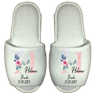 May include: Pair of white slippers with a pink floral design and the letter H. The slippers are personalised with the name Helena, the word Bride, and the date 21.05.2021.