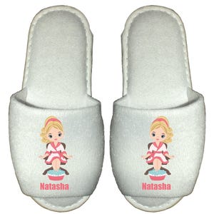A pair of white spa slippers featuring a cartoon illustration of a girl in a robe with her feet in a bowl of water. The slippers are personalised with a name printed on them.