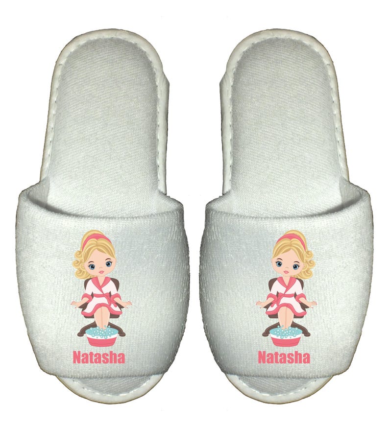 A pair of white spa slippers featuring a cartoon illustration of a girl in a robe with her feet in a bowl of water. The slippers are personalised with a name printed on them.