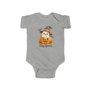 May include: A heather gray short-sleeve bodysuit featuring a graphic of a white duckling wearing a brown witch hat, sitting inside an orange jack-o'-lantern pumpkin. The design includes small bats and stars, with the text Tiny Spook printed below.