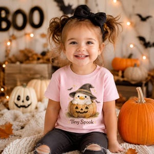 May include: A light pink short-sleeved t-shirt featuring a graphic of a white duck wearing a black witch hat and sitting inside a carved jack-o'-lantern pumpkin. The text Tiny Spook is printed in brown below the pumpkin illustration.