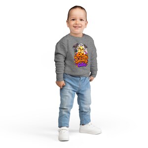 May include: A heather gray long-sleeve sweatshirt featuring a graphic of a yellow chick wearing a ghost costume, sitting inside a carved orange jack-o'-lantern, surrounded by bats and the text Spooky Cutie in purple and orange lettering.