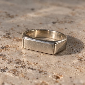 Puede incluir: Un anillo de metal plateado con una parte superior rectangular y plana. La banda es lisa y sólida, con un acabado pulido que refleja la luz en toda su superficie. El diseño es sencillo y uniforme, mostrando una forma geométrica limpia.