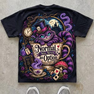 May include: A black t-shirt featuring a vibrant graphic of a purple striped cat in a teacup. The text reads "Normal Was Never an Option". The design includes a top hat, a pocket watch, a "Drink Me" bottle, a book, mushrooms, and playing cards.
