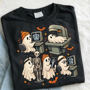 May include: A black t-shirt featuring a graphic print of ghosts dressed as medical professionals. The design includes ghosts operating computers, examining x-rays of a skeleton, and using medical equipment, accented with orange bats and stars.