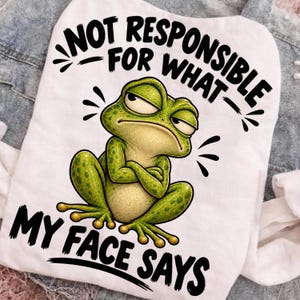 May include: A white t-shirt featuring a graphic of a green frog with a grumpy expression and crossed arms. The text above the frog reads NOT RESPONSIBLE FOR WHAT and the text below the frog reads MY FACE SAYS in bold black lettering.