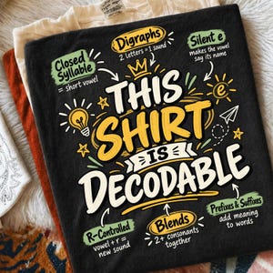May include: A black t-shirt featuring a graphic with the text "THIS SHIRT IS DECODABLE" in yellow and white. The design includes educational annotations like "Digraphs," "Silent e," "Closed Syllable," "R-Controlled," "Blends," and "Prefixes & Suffixes."