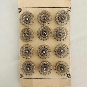 May include: A set of twelve circular, clear glass buttons arranged in a four-by-three grid on a rectangular beige cardboard backing. Each button features a faceted center point surrounded by a textured, radial pattern with small raised dots.