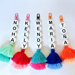 May include: Five personalized keychains featuring white circular beads with black letters spelling NOHA, KELLY, WENDY, MARIA, and TOÑA. Each keychain has a colorful layered tassel at the bottom and a metal lobster clasp in pink, black, or orange.