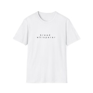 May include: A white short-sleeved t-shirt featuring the text "bread whisperer" printed in small, black, lowercase sans-serif letters centered on the chest. The garment has a classic crew neckline and a standard, straight-cut silhouette.