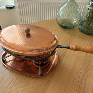 Op de afbeelding: Een ovale serveerschaal van gehamerd koper met een bijpassend deksel met houten knop. De schaal rust op een zwarte metalen standaard boven een koperen onderblad en heeft een lange zwarte metalen handgreep met een houten greep voor transport.