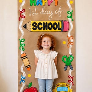 May include: A vertical brown paper banner featuring the text HAPPY 1st day of SCHOOL in colorful letters. The banner is decorated with hand-drawn illustrations of a yellow pencil, an orange crayon, green scissors, a red apple, and a yellow school bus.
