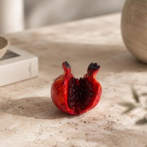 Könnte beinhalten: Eine dekorative Granatapfel-Skulptur mit einer leuchtend roten Außenseite und einer strukturierten, rissigen Oberfläche. Die Frucht ist aufgebrochen und zeigt dunkle, glänzende rote Kerne sowie zwei markante, verkohlte Stiele oben.