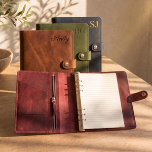 Puede incluir: Una colección de cuatro agendas de cuero con cierres a presión. Tres están cerradas y apiladas, con los nombres Andy, BG y SJ. La cuarta está abierta, mostrando una cubierta color granate, un bolsillo con cremallera y un archivador de seis anillas.