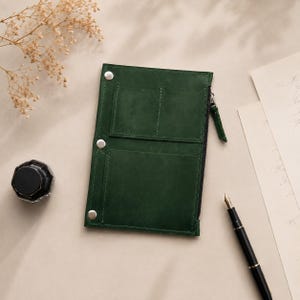 May include: A rectangular dark green leather organiser featuring two top pockets, one larger bottom pocket, and a side zip closure. The organiser is secured with three silver-toned metal snaps along the left edge, providing a structured design for holding small items.