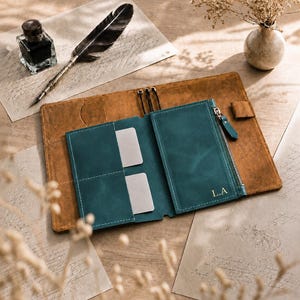 Peut inclure: Une couverture de journal en cuir beige avec un insert en cuir bleu sarcelle comprenant des fentes pour cartes et une poche zippée. L'insert bleu sarcelle porte les initiales L.A. en lettres dorées. La couverture inclut des cordons élastiques.