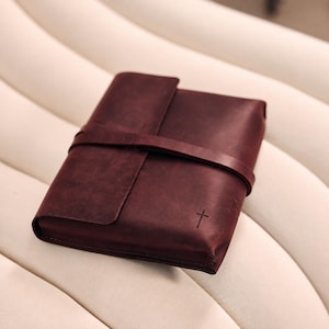 Peut inclure: Une couverture de journal en cuir bordeaux foncé avec une finition mate lisse. La couverture présente une fermeture à sangle en cuir enveloppante et un petit symbole de croix simple gaufré dans le coin inférieur droit du rabat avant.