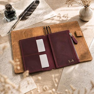 Puede incluir: Una funda de cuero color canela para cuaderno con un inserto de cuero color burdeos oscuro adjunto, que incluye dos ranuras para tarjetas y un bolsillo con cremallera. El inserto burdeos tiene grabadas las iniciales L.A. y un tirador de cremallera plateado.