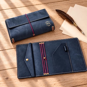 Peut inclure: Une couverture de journal en cuir bleu foncé avec des coutures blanches, les lettres WA dorées en relief, une fermeture élastique rose, et un système élastique intérieur rose assorti avec un passant pour stylo et une poche triangulaire à bouton-pression doré.