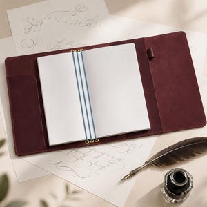 May include: A deep burgundy leather notebook cover featuring a refillable insert held by three vertical blue elastic bands with brass eyelets. The open cover includes a pen loop on the right side, accompanied by a dark feather quill and an inkwell.