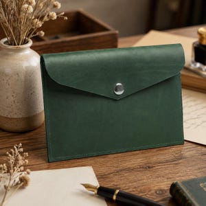 Pode incluir: Um porta-documentos de couro verde-floresta estilo envelope com acabamento fosco liso, aba pontiaguda e um fecho de botão de pressão de metal prateado. O estojo retangular é costurado ao longo das bordas com linha verde correspondente.