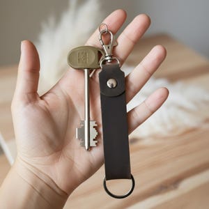 May include: A dark brown leather keychain strap with a silver metal lobster clasp and a split ring, attached to a long silver key featuring an olive green plastic head with a geometric logo embossed on the surface of the key handle.