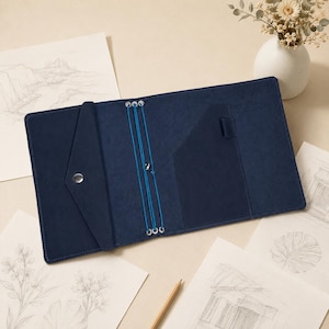 May include: A dark blue leather traveler's notebook cover featuring a triangular pocket with a silver snap button, a vertical pocket, and three bright blue elastic cords held by silver eyelets for securing multiple notebook inserts.