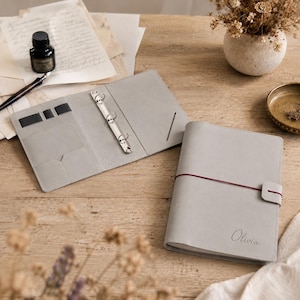 May include: A light grey leather binder with a three-ring metal mechanism, interior card slots, and pen loops. A closed version of the same binder features a dark red elastic closure strap and the name Olivia embossed on the front cover.