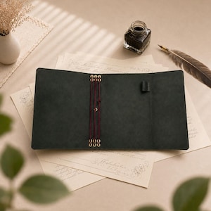 Puede incluir: Una funda para cuaderno de cuero gris carbón oscuro con tres cordones elásticos verticales de color burdeos asegurados con ojales metálicos dorados. El interior incluye una pequeña trabilla de cuero negro para bolígrafo.