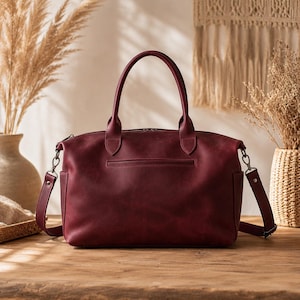 Peut inclure: Un sac à main en cuir bordeaux profond doté de deux poignées supérieures, d'une poche zippée horizontale sur le devant et d'une bandoulière amovible avec des attaches en métal argenté fixées sur les côtés du sac.
