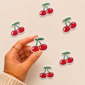 Puede incluir: Seis parches bordados de cerezas que muestran dos cerezas de color rojo brillante con tallos y hojas verdes. Cada parche tiene un borde blanco y un acabado de hilo texturizado. Están dispuestos de forma dispersa con uno sostenido al frente.