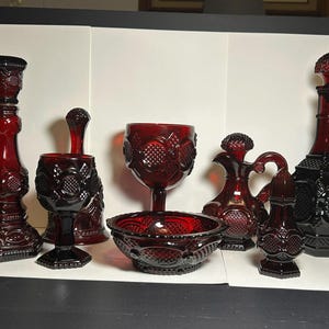 May include: A collection of seven ruby red pressed glass items featuring a repeating geometric hobnail and diamond pattern. The set includes a tall candlestick, two stemmed goblets, a small bowl, a creamer pitcher, a shaker, and a decanter.