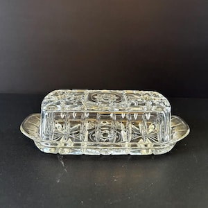 May include: A clear glass butter dish featuring a rectangular lid and base with a pressed geometric starburst pattern. The lid has a flat top with a central star motif and vertical fluted sides, while the base has a scalloped edge design.