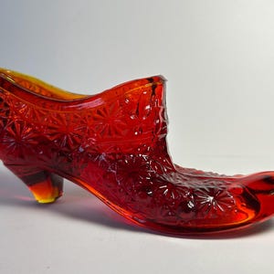 May include: A translucent red glass shoe figurine featuring a pressed starburst pattern across the exterior surface. The decorative object has a pointed toe, a raised heel, and a smooth, curved opening at the top, showcasing a vibrant ruby hue.